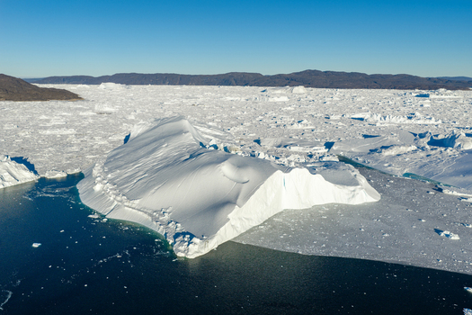 Aerial view of Ilulissat glacier featuring a massive iceberg floating in dark blue waters surrounded by a vast expanse of sea ice and distant rocky coastline in Greenland. Aerial view of Ilulissat glacier featuring a massive iceberg floating in dark blue waters surrounded by a vast expanse of sea ice and distant rocky coastline in Greenland.