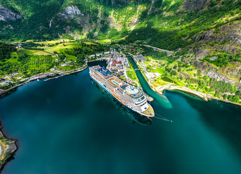 Aerial view of a large cruise ship docked in the fjord at Flam surrounded by steep green mountains lush hills and small buildings in Flam Vestland Norway. Aerial view of a large cruise ship docked in the fjord at Flam surrounded by steep green mountains lush hills and small buildings in Flam Vestland Norway.