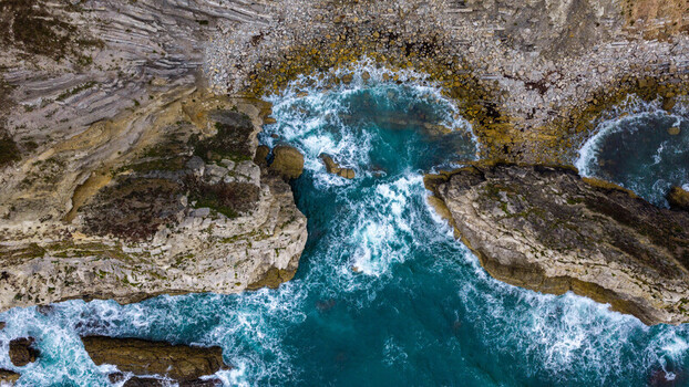Aerial view of Lulworth Cove showing turquoise sea water crashing against rugged limestone cliffs and rocky coastline in Wareham England United Kingdom. Aerial view of Lulworth Cove showing turquoise sea water crashing against rugged limestone cliffs and rocky coastline in Wareham England United Kingdom.