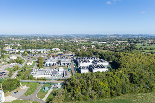 Aerial view of a data center complex surrounded by lush green trees and fields in the United Kingdom.