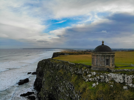 Aerial view of Mussenden Temple perched on the edge of dramatic cliffs overlooking the Atlantic Ocean with green fields and a cloudy sky in Coleraine Northern Ireland United Kingdom. Aerial view of Mussenden Temple perched on the edge of dramatic cliffs overlooking the Atlantic Ocean with green fields and a cloudy sky in Coleraine Northern Ireland United Kingdom.