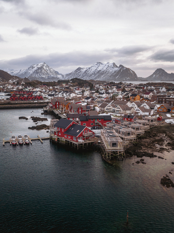 Aerial view of red cabins clustered along the rugged coastline contrasting with the dark rippled waters and snow dusted peaks beyond Ballstad Nordland Norway. Aerial view of red cabins clustered along the rugged coastline contrasting with the dark rippled waters and snow dusted peaks beyond Ballstad Nordland Norway.