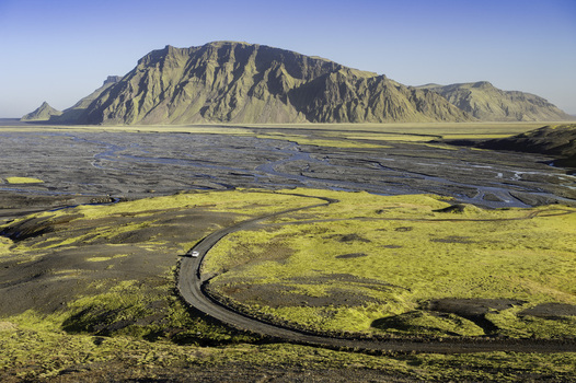 Katla Geopark Iceland 21 October 2025 View of a winding road through mossy hills with a white car and a massive mountain in the background. Katla Geopark Iceland 21 October 2025 View of a winding road through mossy hills with a white car and a massive mountain in the background.