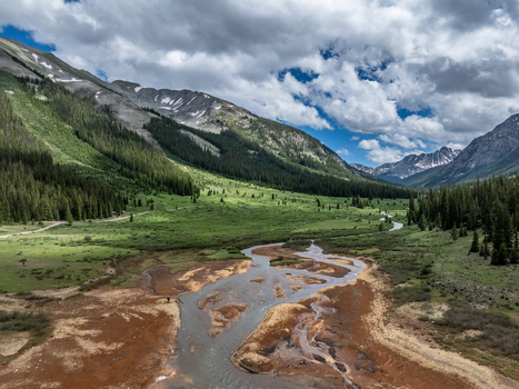 View of a winding river flowing through a lush green mountain valley with dense pine forests and rocky peaks under a dramatic cloudy sky Colorado United States.