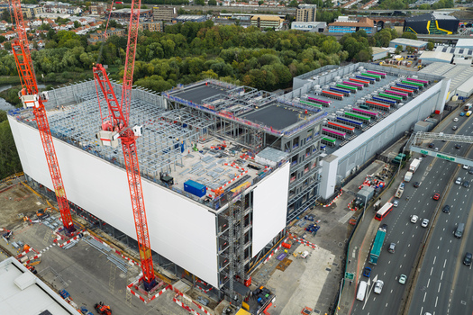 Aerial view of a large data center construction site featuring tall red cranes steel framework and colorful cooling units on the roof situated next to a busy highway in the United Kingdom.