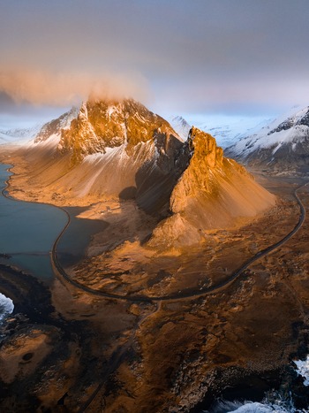 Aerial view of Eystrahorn mountain illuminated by golden hour light with snow capped peaks a winding coastal road and the Atlantic Ocean in Sveitarfelagid Hornafjordur Iceland. Aerial view of Eystrahorn mountain illuminated by golden hour light with snow capped peaks a winding coastal road and the Atlantic Ocean in Sveitarfelagid Hornafjordur Iceland.