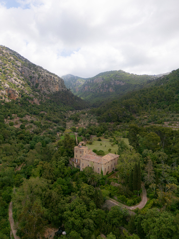 Aerial view of a historic stone estate nestled among lush green forests and rugged mountains under a cloudy sky in Valldemossa Balearic Islands Spain.
