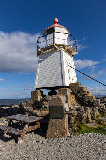 View of the Laukvik fyr lighthouse standing on a rocky shore with a wooden picnic table under a blue sky in Laukvik Nordland Norway. View of the Laukvik fyr lighthouse standing on a rocky shore with a wooden picnic table under a blue sky in Laukvik Nordland Norway.