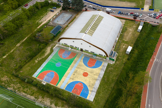 Aerial view of a modern sports hall with a large arched roof and vibrant outdoor basketball courts surrounded by green trees and urban infrastructure San Giovanni Valdarno Tuscany Italy.