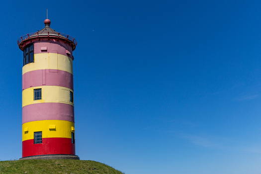 View of the iconic Pilsum lighthouse with its distinctive red and yellow stripes standing on a grassy mound under a clear blue sky in Krummhorn Niedersachsen Germany. View of the iconic Pilsum lighthouse with its distinctive red and yellow stripes standing on a grassy mound under a clear blue sky in Krummhorn Niedersachsen Germany.