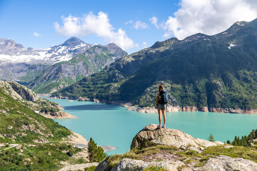 View of a hiker standing on a rock overlooking the turquoise waters of Emosson Lake surrounded by majestic snow capped mountains in Le Chatelard Valais Switzerland. View of a hiker standing on a rock overlooking the turquoise waters of Emosson Lake surrounded by majestic snow capped mountains in Le Chatelard Valais Switzerland.