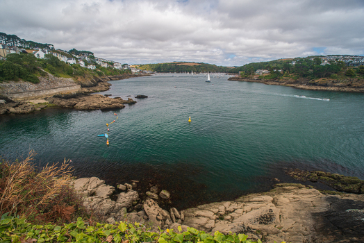 View of the Fowey River with boats sailing towards the town of Fowey featuring rocky foreground cliffs and a cloudy sky in Fowey Cornwall United Kingdom. View of the Fowey River with boats sailing towards the town of Fowey featuring rocky foreground cliffs and a cloudy sky in Fowey Cornwall United Kingdom.