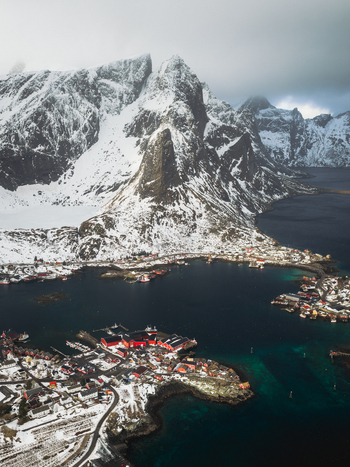 Aerial view of Reinebringen s craggy snow dusted peaks overlooking a tranquil bay dotted with vibrant red roofed buildings and fishing boats Reinebringen Nordland Norway.