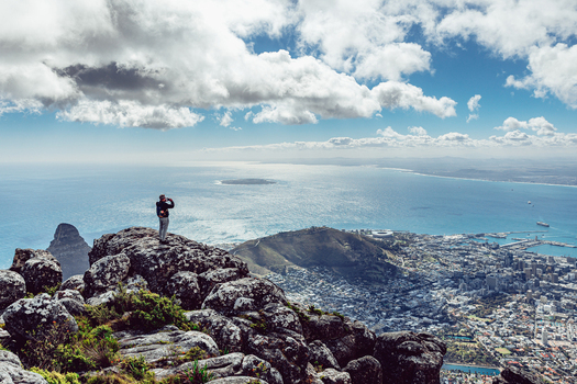 View of a person standing on a rocky mountain peak overlooking the city coastline and ocean under a cloudy sky in Cape Town Western Cape South Africa. View of a person standing on a rocky mountain peak overlooking the city coastline and ocean under a cloudy sky in Cape Town Western Cape South Africa.