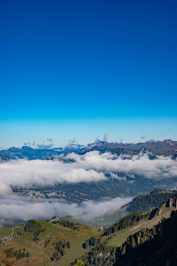 Aerial view of the Swiss Alps with rolling green slopes scattered trees and a layer of clouds resting in the valley under a clear blue sky in Airolo Ticino Switzerland. Aerial view of the Swiss Alps with rolling green slopes scattered trees and a layer of clouds resting in the valley under a clear blue sky in Airolo Ticino Switzerland.