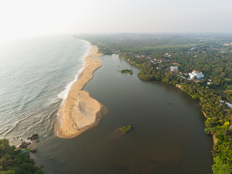 Aerial view of a narrow sandbar separating the turquoise ocean waves from a calm lagoon surrounded by lush tropical palm trees and coastal buildings in Galle Southern Province Sri Lanka.