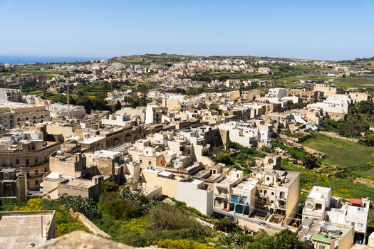 View of traditional limestone buildings and flat rooftops overlooking green fields and the Mediterranean Sea under a clear blue sky in Zebbug Gozo Region Malta. View of traditional limestone buildings and flat rooftops overlooking green fields and the Mediterranean Sea under a clear blue sky in Zebbug Gozo Region Malta.