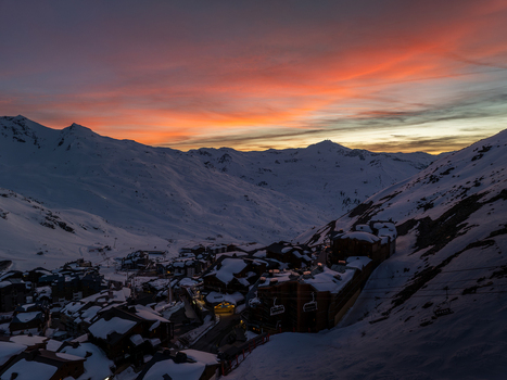 Aerial view of snow blanketed chalets nestled in a valley the sky ablaze with fiery oranges and soft pinks against the stark white peaks Val Thorens Auvergne Rh&ocirc;ne Alpes France.