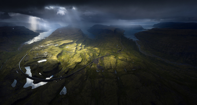 Aerial view of a winding road through green mossy hills under dramatic sunbeams and dark storm clouds in Iceland.