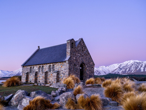 View of the Church of the Good Shepherd a historic stone church set against snow capped mountains under a purple sky with tussock grasses Lake Tekapo Canterbury Region New Zealand.