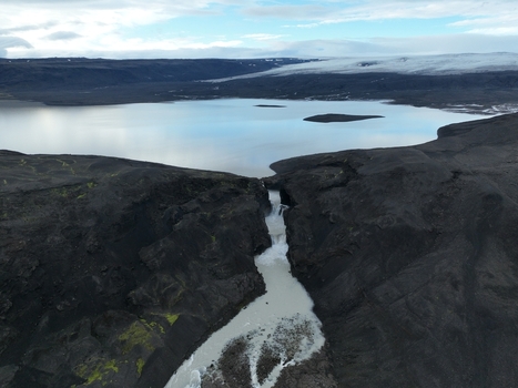 Aerial view of Nyifoss waterfall flowing from a glacial lake through a rugged volcanic canyon with dark basalt rocks and distant ice fields in Reykholt Borgarbyggd Iceland.