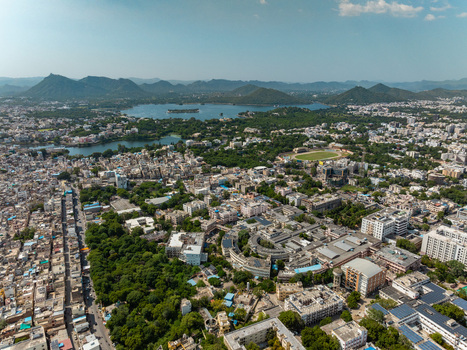 Aerial view of the city of Udaipur featuring dense urban architecture lush green trees and the expansive Pichola Lake in the background surrounded by hills. Udaipur Rajasthan India.