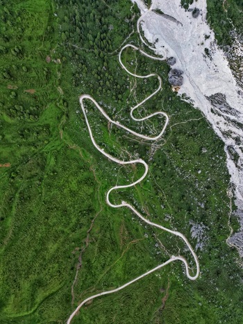 Aerial view of the winding mountain road through lush green slopes and rocky terrain in the Dolomite Mountains Canazei Trentino South Tyrol Italy. Aerial view of the winding mountain road through lush green slopes and rocky terrain in the Dolomite Mountains Canazei Trentino South Tyrol Italy.