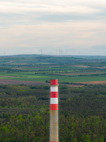 Aerial view of a red and white industrial chimney rising above a dense forest with wind turbines scattered across the distant agricultural fields in Breclav South Moravian Region Czechia.