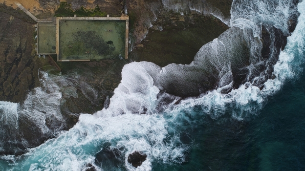Aerial view of Newport Rock Pool with crashing white waves against dark coastal rocks and swimmers in the rectangular ocean pool in Sydney New South Wales Australia.