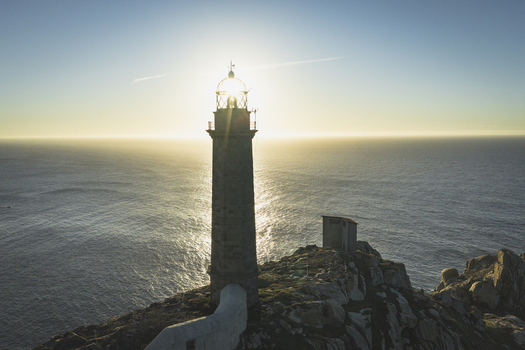 Aerial view of a stone lighthouse perched on a rocky cliff overlooking the Atlantic Ocean at sunset with golden light reflecting on the water in Galicia Spain.