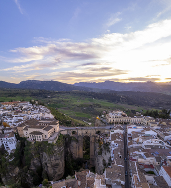 Aerial view of the historic Puente Nuevo bridge spanning the deep El Tajo gorge surrounded by white buildings and rugged mountains under a soft sunset sky in Ronda Andalusia Spain. Aerial view of the historic Puente Nuevo bridge spanning the deep El Tajo gorge surrounded by white buildings and rugged mountains under a soft sunset sky in Ronda Andalusia Spain.