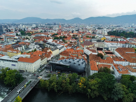 Aerial view of the Kunsthaus Graz with its unique organic architecture surrounded by historic red tiled rooftops the Mur river and urban infrastructure in Graz Styria Austria.