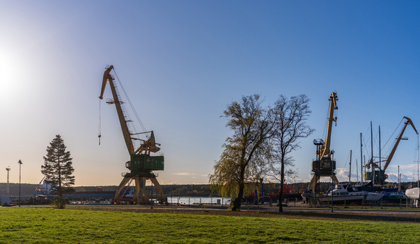 View of industrial port cranes standing along the harbor with boats moored at the dock surrounded by green grass and trees under a clear blue sky in Klaipeda Klaipeda County Lithuania. View of industrial port cranes standing along the harbor with boats moored at the dock surrounded by green grass and trees under a clear blue sky in Klaipeda Klaipeda County Lithuania.
