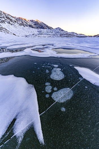 View of methane bubbles trapped in the frozen surface of a lake with snow covered mountains under a clear sky in Madesimo Lombardy Italy. View of methane bubbles trapped in the frozen surface of a lake with snow covered mountains under a clear sky in Madesimo Lombardy Italy.