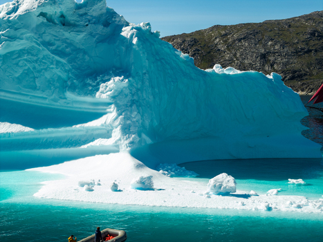 Aerial view of a massive iceberg floating in turquoise waters with a small boat carrying people nearby set against a rugged rocky coastline in Greenland. Aerial view of a massive iceberg floating in turquoise waters with a small boat carrying people nearby set against a rugged rocky coastline in Greenland.