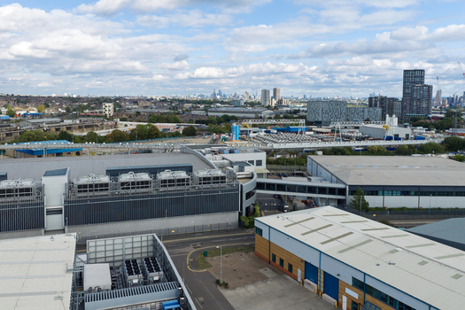Aerial view of a large data center facility featuring rooftop cooling units industrial buildings and a connecting bridge with a distant city skyline under a cloudy sky in the United Kingdom. Aerial view of a large data center facility featuring rooftop cooling units industrial buildings and a connecting bridge with a distant city skyline under a cloudy sky in the United Kingdom.