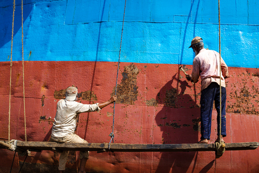 Keraniganj Bangladesh 31 August 2024 View of two workers on a wooden scaffolding scraping rust from the red and blue hull of a large vessel at the Shipyard Dhaka.
