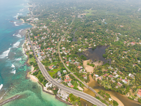 Aerial view of the coastline featuring Devil s Rock lush tropical vegetation and residential buildings along the road in Ahangama Southern Province Sri Lanka. Aerial view of the coastline featuring Devil s Rock lush tropical vegetation and residential buildings along the road in Ahangama Southern Province Sri Lanka.