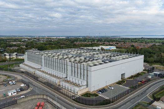 Aerial view of a large data center facility with cooling units on the roof surrounded by roads and parking areas in the United Kingdom.