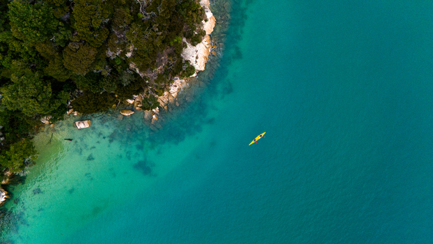 Aerial view of a lone yellow kayak gliding across the turquoise waters near a rocky tree lined coast Abel Tasman National Park New Zealand. Aerial view of a lone yellow kayak gliding across the turquoise waters near a rocky tree lined coast Abel Tasman National Park New Zealand.