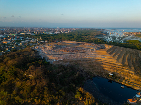 Aerial view of TPA Suwung landfill with terraced slopes and waste management areas under golden hour light near urban sprawl in Kecamatan Denpasar Selatan Bali Indonesia.