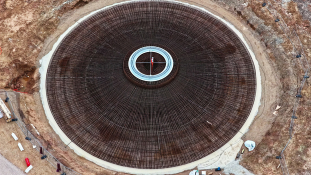 Aerial view of a circular wind turbine foundation under construction featuring an intricate steel rebar reinforcement mesh and a central metal ring set within an excavated site in Latvia. Aerial view of a circular wind turbine foundation under construction featuring an intricate steel rebar reinforcement mesh and a central metal ring set within an excavated site in Latvia.