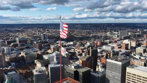 Aerial view of the Baltimore city skyline featuring a prominent American flag waving in the foreground against a cloudy sky showcasing urban architecture in Baltimore Maryland United States.