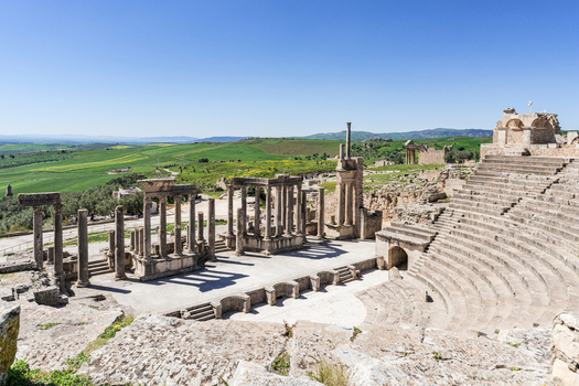 View of the ancient Roman theater at Dougga Archaeological Site with stone columns and tiered seating set against green hills under a clear blue sky in Teboursouk Beja Governorate Tunisia.