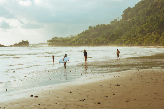 View of people on a sandy beach with a surfboard lush green hills and ocean waves under soft golden hour light in Costa Rica.