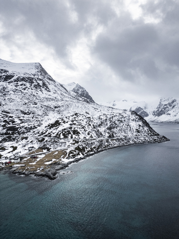 Aerial view of Grotfjord with snow covered mountains a winding coastal road and dark blue waters under a cloudy sky in Tromvik Troms Norway. Aerial view of Grotfjord with snow covered mountains a winding coastal road and dark blue waters under a cloudy sky in Tromvik Troms Norway.
