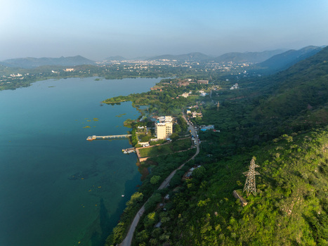 Aerial view of a lake surrounded by lush green hills and a building on the shore with a power line tower on the hillside in Udaipur Rajasthan India. Aerial view of a lake surrounded by lush green hills and a building on the shore with a power line tower on the hillside in Udaipur Rajasthan India.