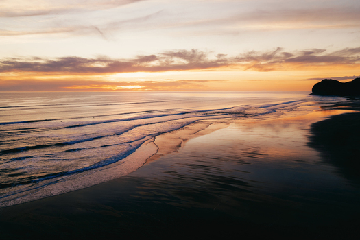 Aerial view of Piha Beach at sunset with gentle waves rolling onto the dark sand reflecting the golden light of the sky Auckland New Zealand. Aerial view of Piha Beach at sunset with gentle waves rolling onto the dark sand reflecting the golden light of the sky Auckland New Zealand.