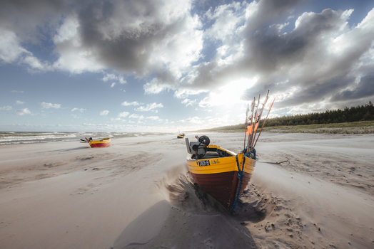 View of traditional fishing boats resting on a sandy beach under a dramatic cloudy sky with sunlight breaking through near a pine forest in Debki Pomeranian Voivodeship Poland. View of traditional fishing boats resting on a sandy beach under a dramatic cloudy sky with sunlight breaking through near a pine forest in Debki Pomeranian Voivodeship Poland.