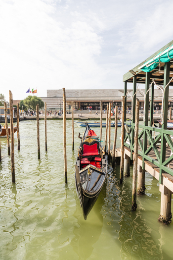 Venice Italy 21 September 2026 View of a traditional black gondola with red velvet seating moored at a wooden pier in the canal with the Santa Lucia station in the background. Venice Italy 21 September 2026 View of a traditional black gondola with red velvet seating moored at a wooden pier in the canal with the Santa Lucia station in the background.